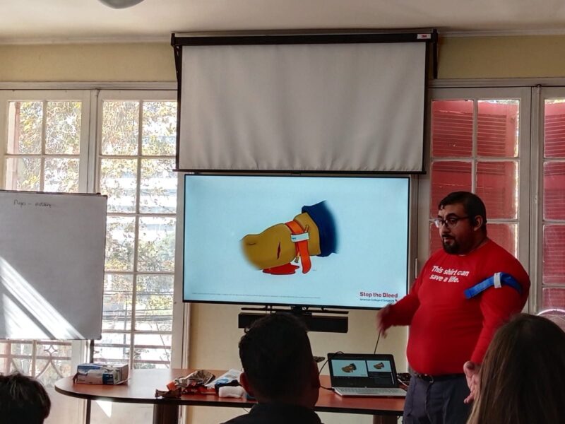 Instructor con polera roja explicando control de hemorragias frente a una pantalla